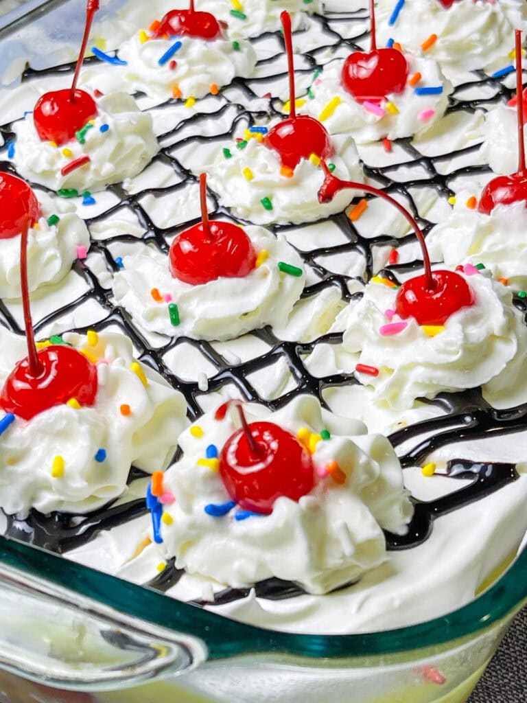 poke cake made with banana cake mix, strawberry jam, vanilla pudding mixed with crushed pineapples and topped with whipped cream and chocolate sauce