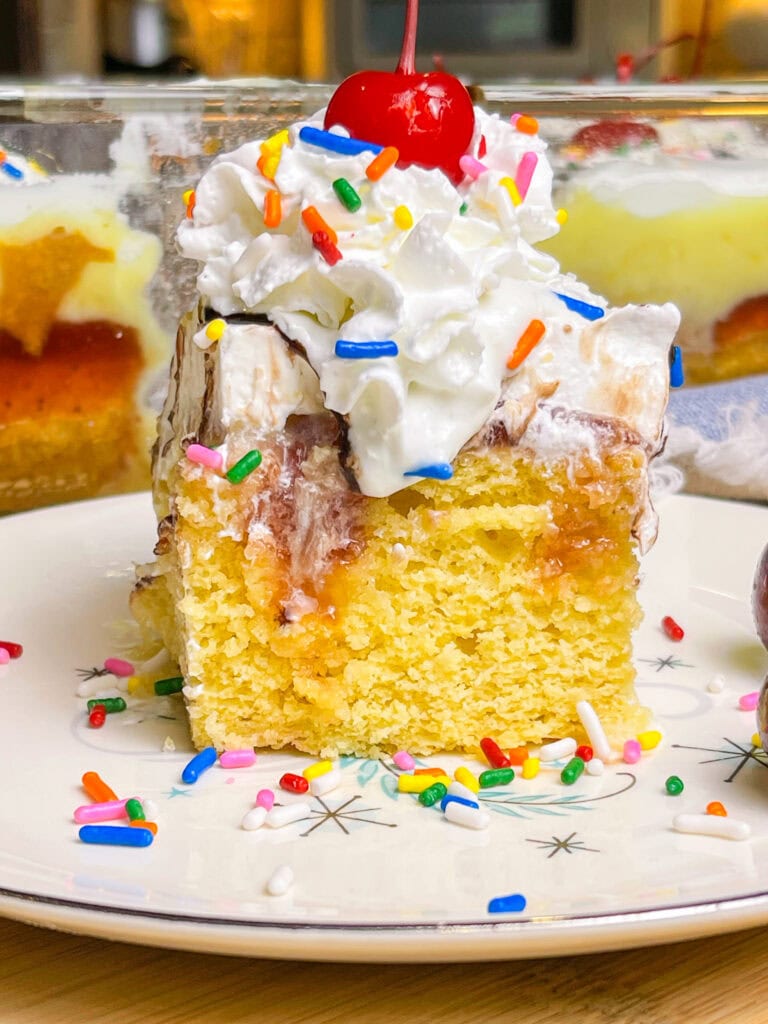 poke cake made with banana cake mix, strawberry jam, vanilla pudding mixed with crushed pineapples and topped with whipped cream and chocolate sauce