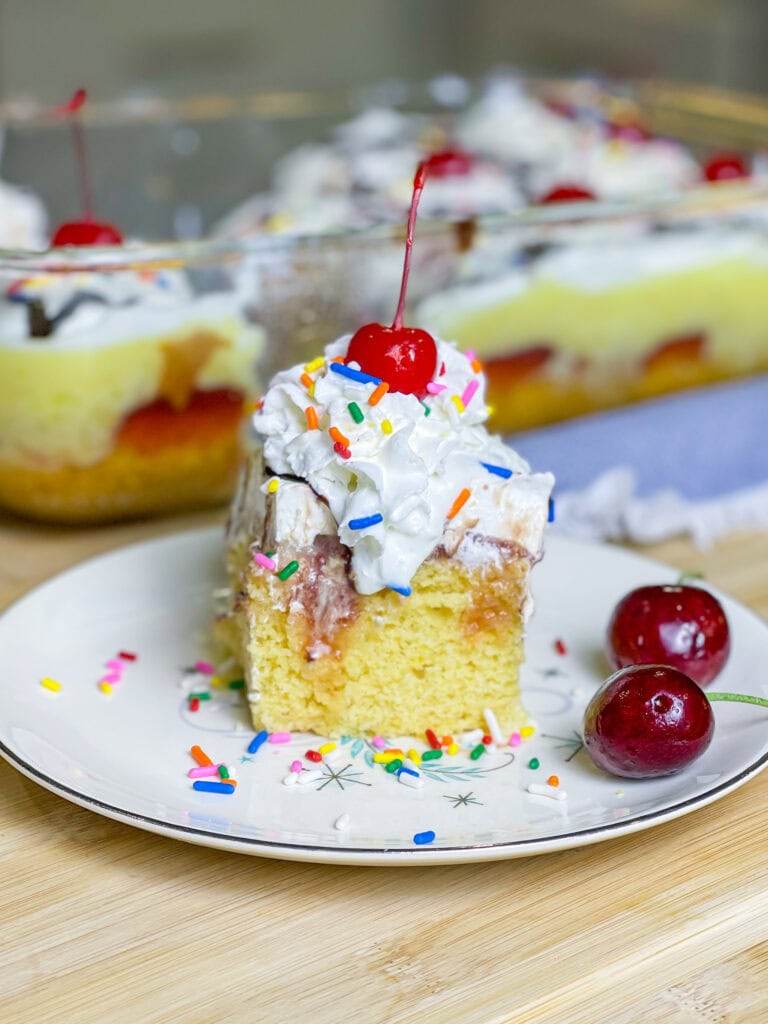 poke cake made with banana cake mix, strawberry jam, vanilla pudding mixed with crushed pineapples and topped with whipped cream and chocolate sauce