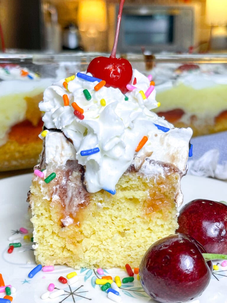 poke cake made with banana cake mix, strawberry jam, vanilla pudding mixed with crushed pineapples and topped with whipped cream and chocolate sauce