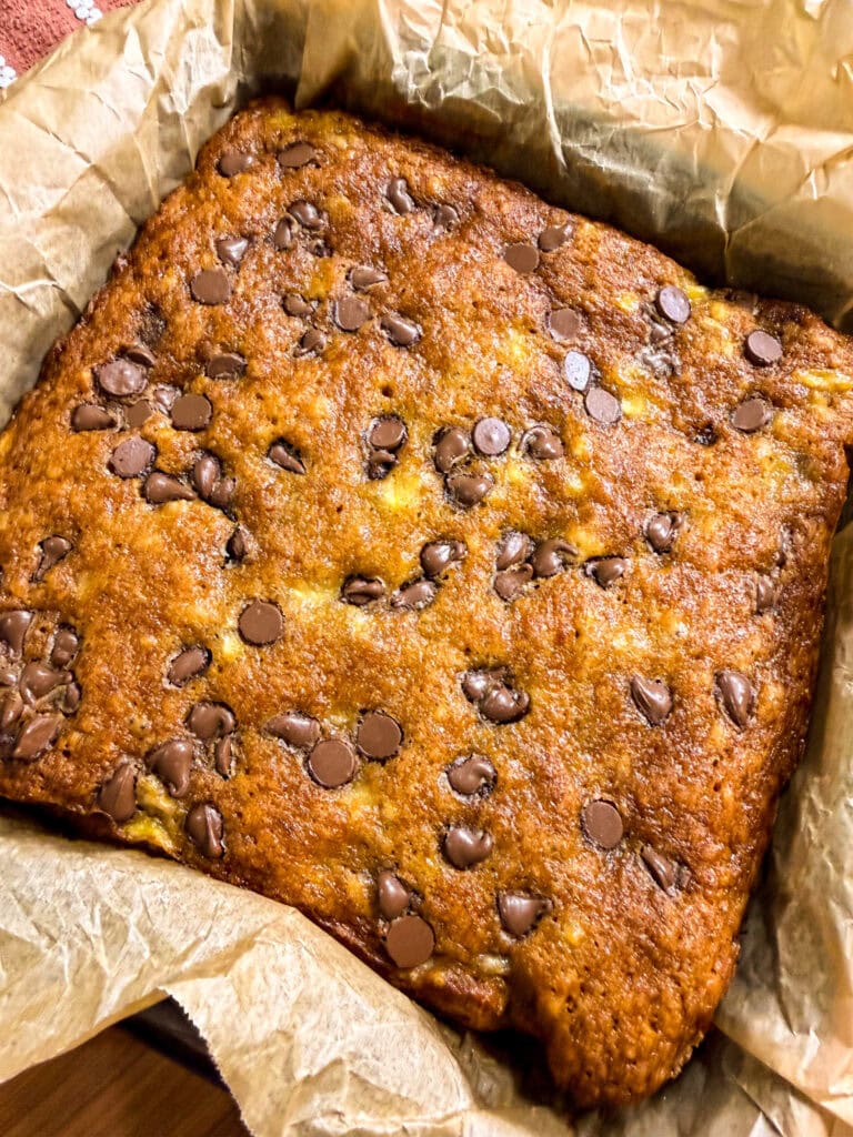 A freshly baked square of Banana Chocolate Chip Cookie Bars sits in a parchment-lined baking pan. The top is golden brown, with melted chocolate chips scattered throughout, reminiscent of classic banana bread but with a delightful cookie bar twist.