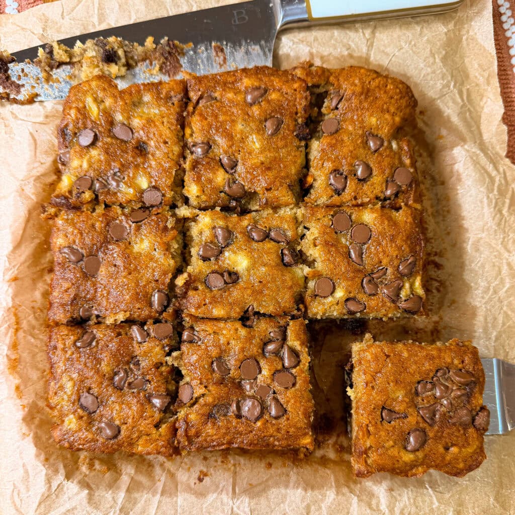 Square pieces of Banana Chocolate Chip Cookie Bars are topped with chocolate chips, then sliced and arranged neatly on parchment paper. A knife and spatula rest beside the bars, ready for serving.