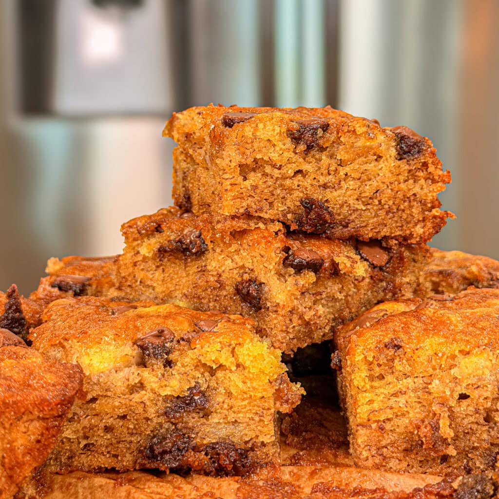 A close-up of several stacked, golden-brown Banana Chocolate Chip Cookie Bars, showing a moist texture and melty chocolate chips, set against a blurred kitchen background.