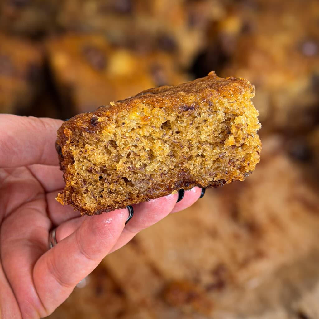 A hand holding a moist, golden-brown square of banana bread with a soft crumb and slightly crisp edges—reminiscent of Banana Chocolate Chip Cookie Bars—with more pieces blurred in the background.