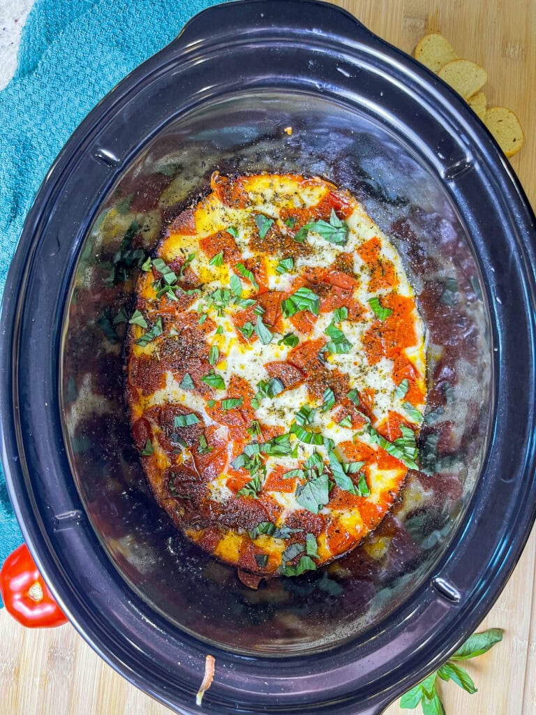 A warm crock pot filled with gooey pepperoni pizza dip, topped with melted cheese and pepperoni slices, served with garlic bagel chips for dipping.