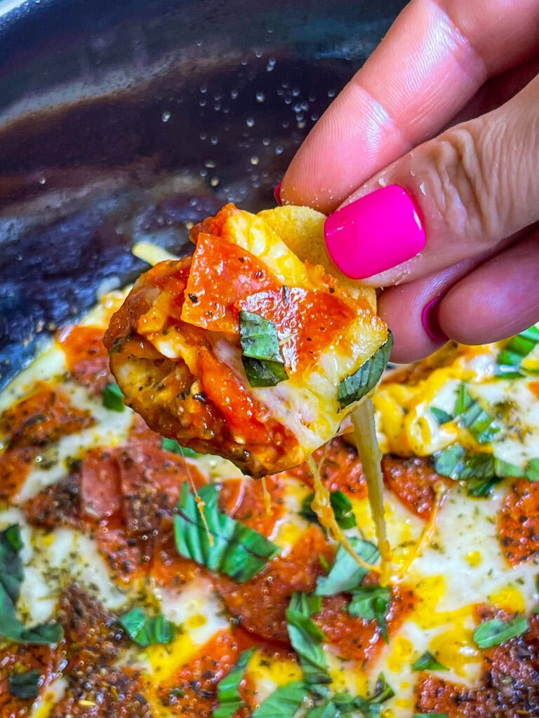A warm crock pot filled with gooey pepperoni pizza dip, topped with melted cheese and pepperoni slices, served with garlic bagel chips for dipping.