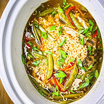 A bowl of slow cooker chicken ramen filled with shredded chicken, ramen noodles, snow peas, carrots, mushrooms, and red peppers, topped with a jammy egg, sesame seeds, and sliced green onions.