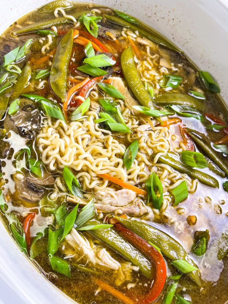 A bowl of slow cooker chicken ramen filled with shredded chicken, ramen noodles, snow peas, carrots, mushrooms, and red peppers, topped with a jammy egg, sesame seeds, and sliced green onions.