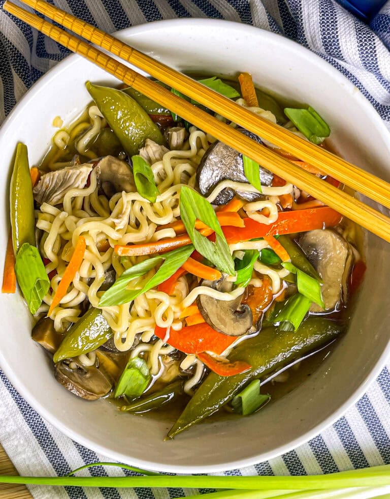 A bowl of slow cooker chicken ramen filled with shredded chicken, ramen noodles, snow peas, carrots, mushrooms, and red peppers, topped with a jammy egg, sesame seeds, and sliced green onions.