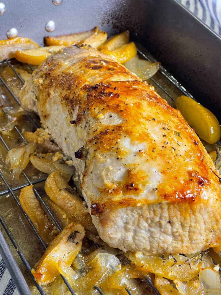 Sliced oven baked apple cider pork loin in a white baking dish surrounded by roasted apples, onions, and a golden cider glaze.