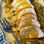 Sliced oven baked apple cider pork loin in a white baking dish surrounded by roasted apples, onions, and a golden cider glaze.