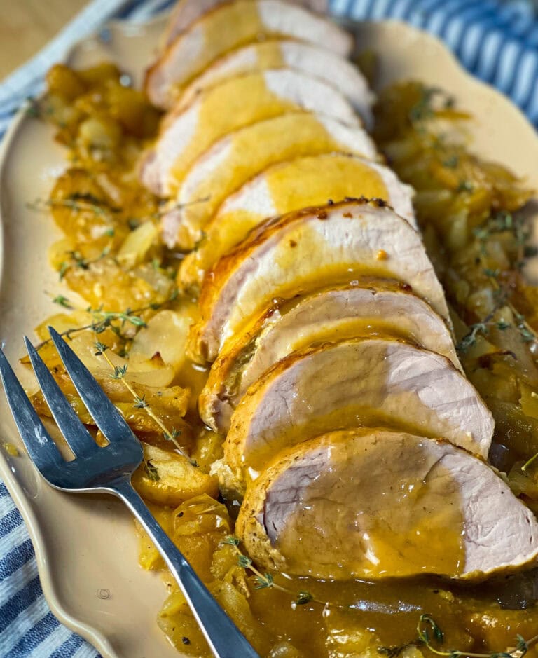 Sliced oven baked apple cider pork loin in a white baking dish surrounded by roasted apples, onions, and a golden cider glaze.