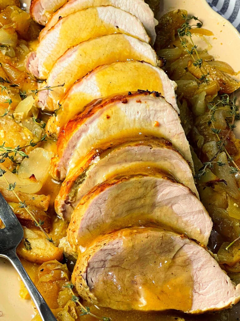 Sliced oven baked apple cider pork loin in a white baking dish surrounded by roasted apples, onions, and a golden cider glaze.