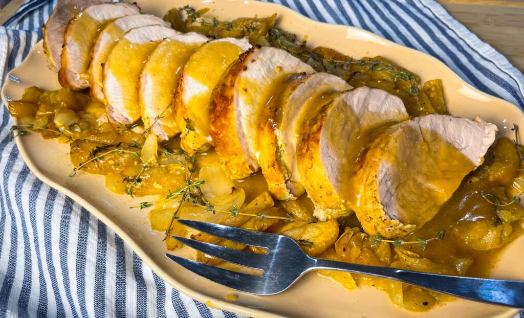 Sliced oven baked apple cider pork loin in a white baking dish surrounded by roasted apples, onions, and a golden cider glaze.
