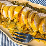 Sliced oven baked apple cider pork loin in a white baking dish surrounded by roasted apples, onions, and a golden cider glaze.