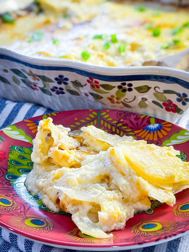 A golden baking dish filled with creamy scalloped potatoes, topped with melted cheddar and fresh green chives, with a serving spoon resting nearby.
