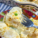 A golden baking dish filled with creamy scalloped potatoes, topped with melted cheddar and fresh green chives, with a serving spoon resting nearby.