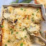 A golden baking dish filled with creamy scalloped potatoes, topped with melted cheddar and fresh green chives, with a serving spoon resting nearby.