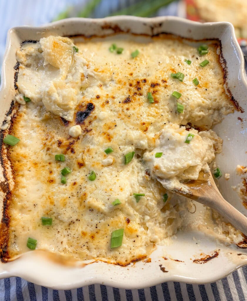 A golden baking dish filled with creamy scalloped potatoes, topped with melted cheddar and fresh green chives, with a serving spoon resting nearby.