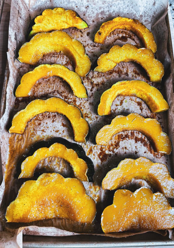 Golden maple roasted acorn squash slices arranged on a parchment-lined baking sheet, glistening with caramelized maple glaze and sprinkled with salted pumpkin seeds.