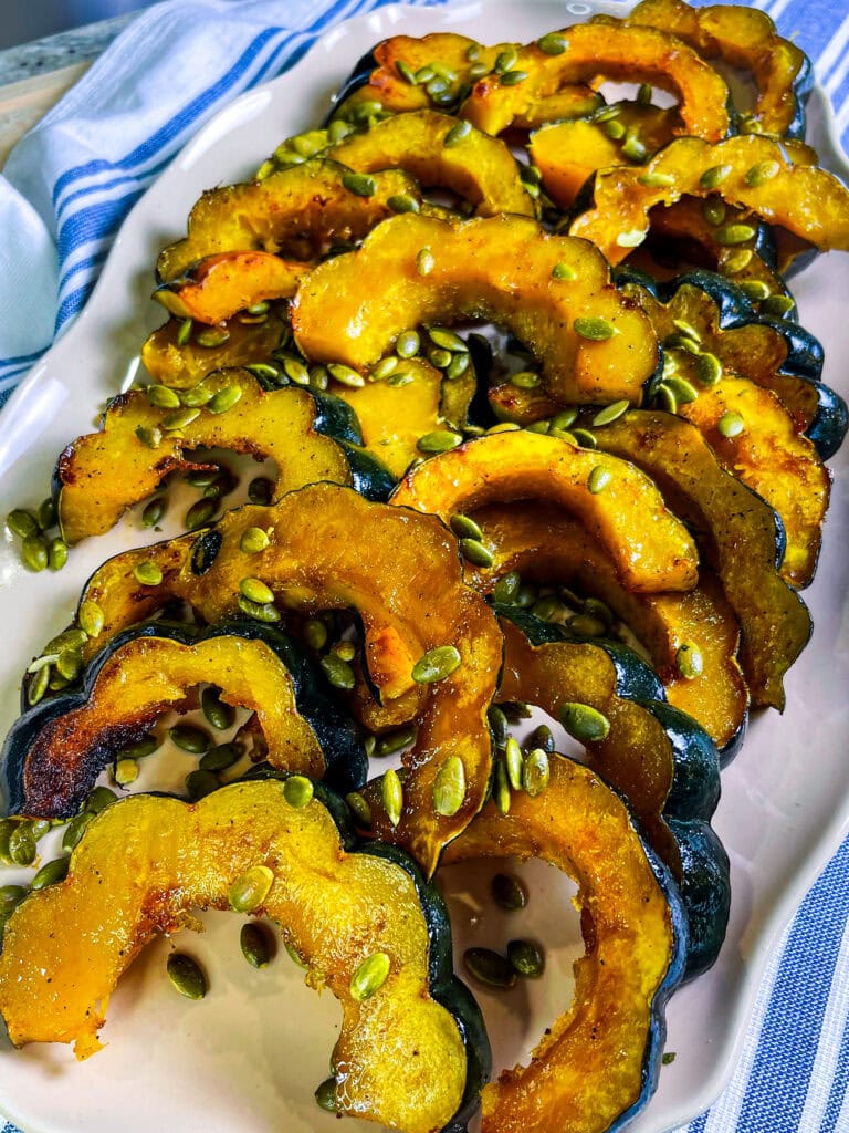 Golden maple roasted acorn squash slices arranged on a parchment-lined baking sheet, glistening with caramelized maple glaze and sprinkled with salted pumpkin seeds.