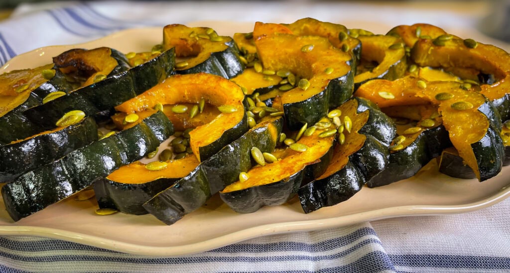 Golden maple roasted acorn squash slices arranged on a parchment-lined baking sheet, glistening with caramelized maple glaze and sprinkled with salted pumpkin seeds.