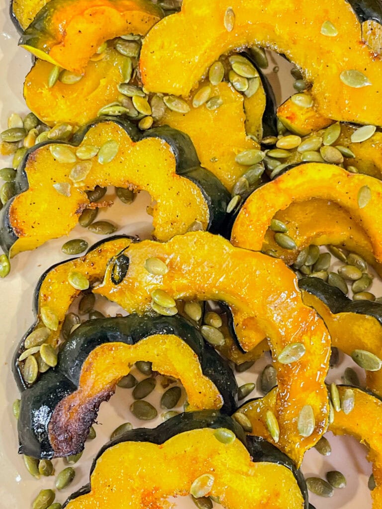 Golden maple roasted acorn squash slices arranged on a parchment-lined baking sheet, glistening with caramelized maple glaze and sprinkled with salted pumpkin seeds.