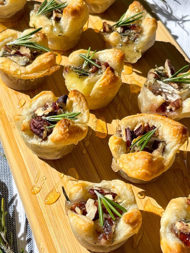 an appetizer of cranberry sauce, brie cheese, rosemary, toasted pecans and honey inside a puff pastry tart lined on a cutting board for serving