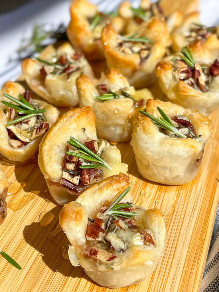 an appetizer of cranberry sauce, brie cheese, rosemary, toasted pecans and honey inside a puff pastry tart lined on a cutting board for serving