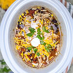 Chicken enchilada soup made with corn, black beans, tomatoes, chicken, and broth in a white crock pot topped with shredded cheddar cheese, cilantro and sour cream