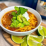 Chicken enchilada soup made with corn, black beans, tomatoes, chicken, and broth in a white crock pot topped with shredded cheddar cheese, cilantro and sour cream
