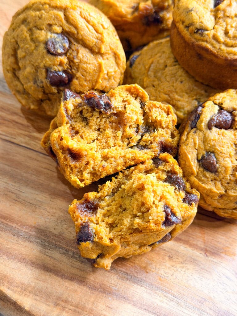 Pumpkin Chocolate Chip Muffins sit invitingly on a wooden surface, with one muffin torn in half to reveal its moist, orange interior studded with rich chocolate chips.