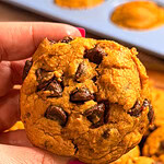 A hand with pink nail polish holds a pumpkin chocolate chip cookie, with more cookies and a blue muffin tin filled with freshly baked Pumpkin Chocolate Chip Muffins in the background.