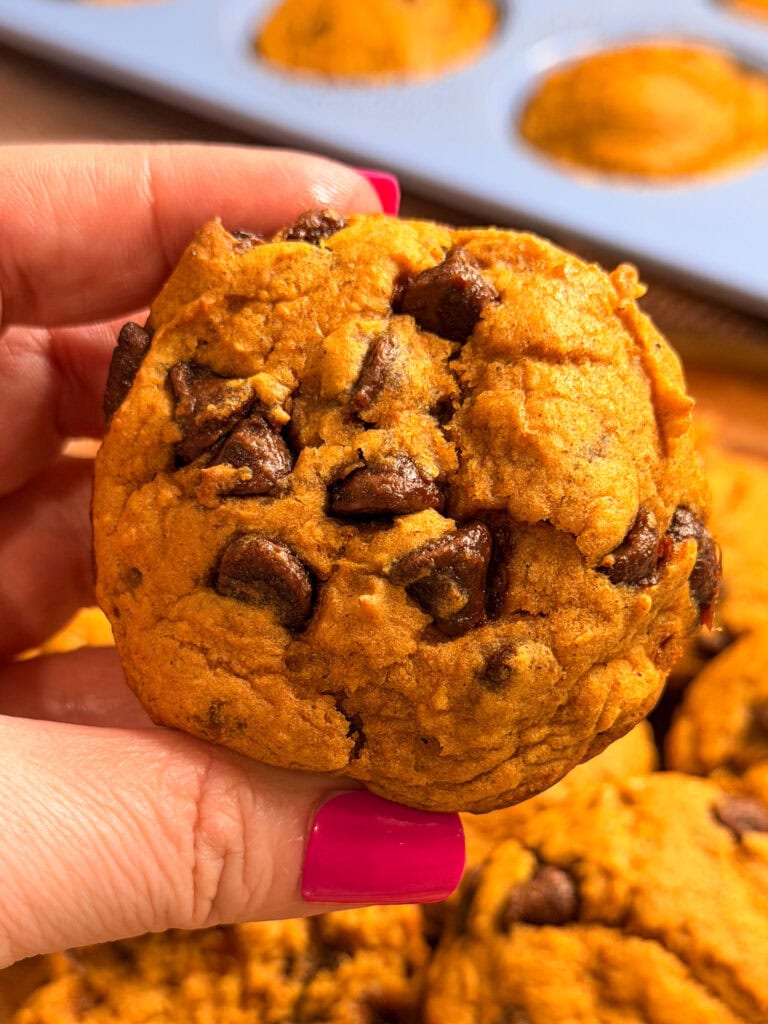 A hand with pink nail polish holds a pumpkin chocolate chip cookie, with more cookies and a blue muffin tin filled with freshly baked Pumpkin Chocolate Chip Muffins in the background.