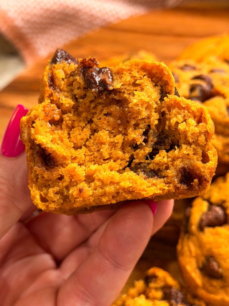 A hand with pink nails holds half of a moist Pumpkin Chocolate Chip Muffin, revealing its soft, textured interior studded with melty chocolate chips. Other muffins are blurred in the background.