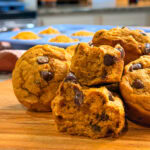 A close-up of Pumpkin Chocolate Chip Muffins on a wooden board, with one muffin split in half showing its moist texture speckled with chocolate chips. More muffins are blurred in the background on a kitchen counter.