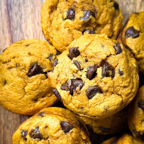 A close-up of Pumpkin Chocolate Chip Muffins arranged on a wooden surface. The muffins are golden-brown with visible chocolate chips.