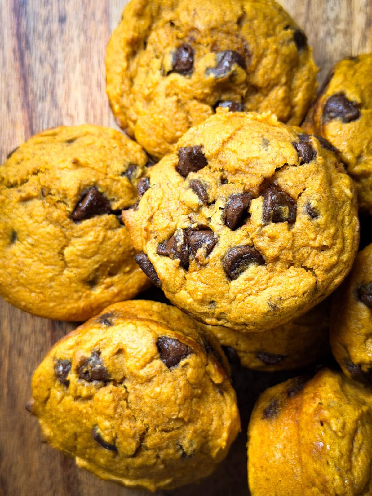A close-up of Pumpkin Chocolate Chip Muffins arranged on a wooden surface. The muffins are golden-brown with visible chocolate chips.