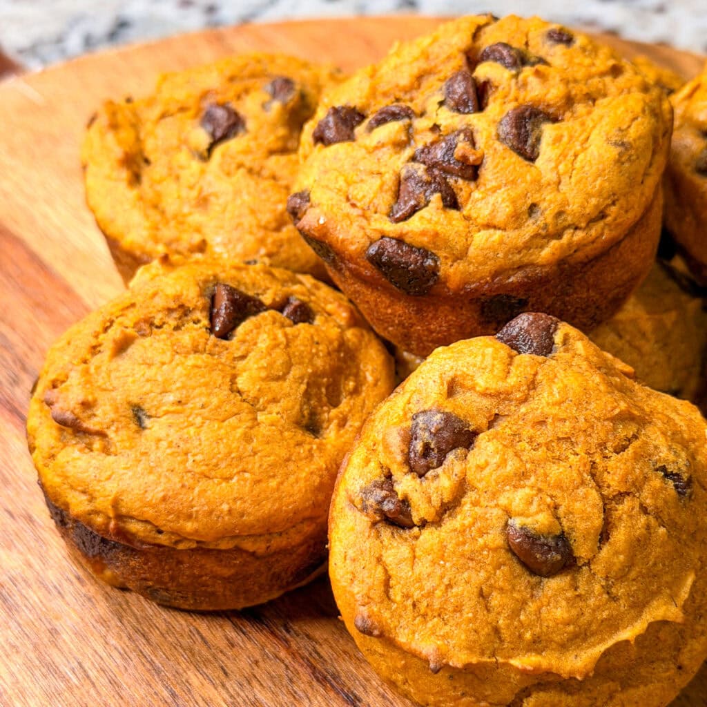 A close-up of several golden-brown Pumpkin Chocolate Chip Muffins, generously studded with chocolate chips on top and arranged on a round wooden serving board.