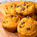 A close-up of several golden-brown Pumpkin Chocolate Chip Muffins, generously studded with chocolate chips on top and arranged on a round wooden serving board.