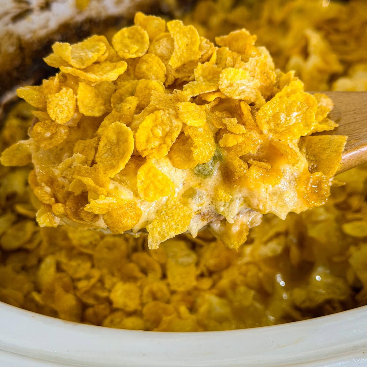 A close-up of a wooden spoon scooping creamy Overnight Hashbrown Breakfast Casserole topped with crunchy cornflakes from a white dish.