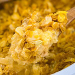 A close-up of a wooden spoon lifting a creamy, cheesy Overnight Hashbrown Breakfast Casserole topped with crunchy cornflakes. The dish appears golden brown with a rich, textured surface.