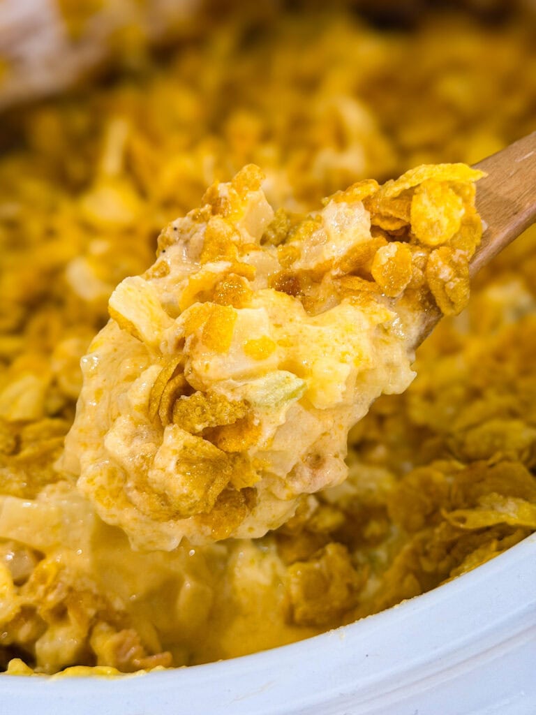 A close-up of a wooden spoon lifting a creamy, cheesy Overnight Hashbrown Breakfast Casserole topped with crunchy cornflakes. The dish appears golden brown with a rich, textured surface.