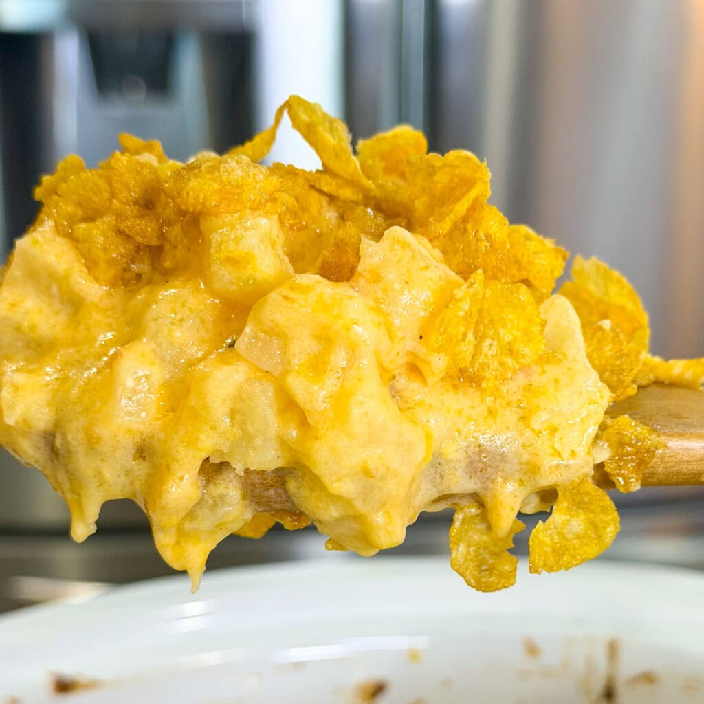 A wooden spoon holds a creamy, cheesy Overnight Hashbrown Breakfast Casserole topped with golden, crunchy cornflakes, with a blurred kitchen background.