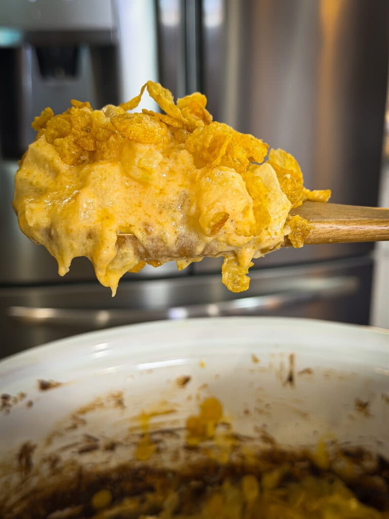 A close-up of a wooden spoon holding a scoop of cheesy Overnight Hashbrown Breakfast Casserole topped with crunchy cornflakes, with a white dish and blurred kitchen appliances in the background.
