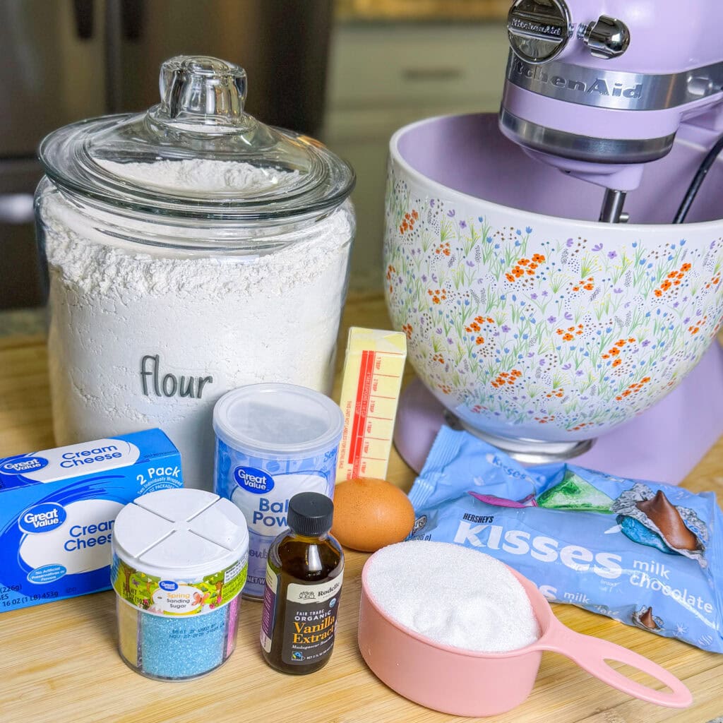 A countertop displays baking ingredients for an Overnight Hashbrown Breakfast Casserole: a jar labeled flour, a stick of butter, an egg, baking powder, vanilla extract, cream cheese, sugar, blue sprinkles, milk chocolate Kisses, and a floral stand mixer.