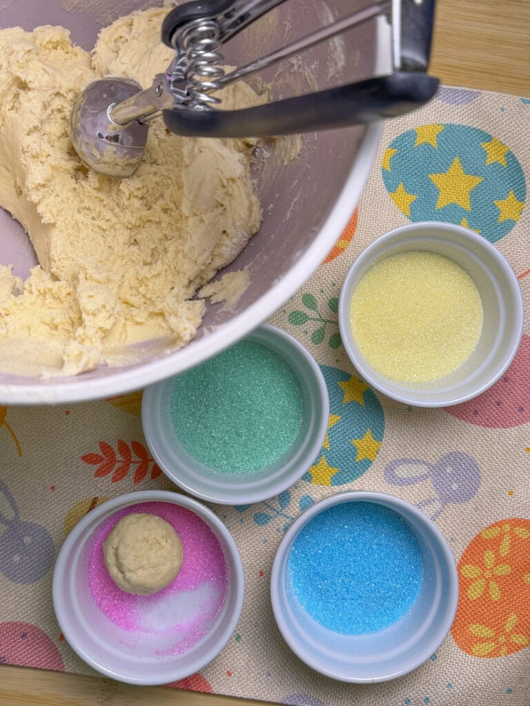 A mixing bowl with cookie dough and a scoop sits beside four small bowls of colored sugar—yellow, green, pink, and blue—on a pastel-patterned mat, evoking the cheerful prep of an Overnight Hashbrown Breakfast Casserole morning.