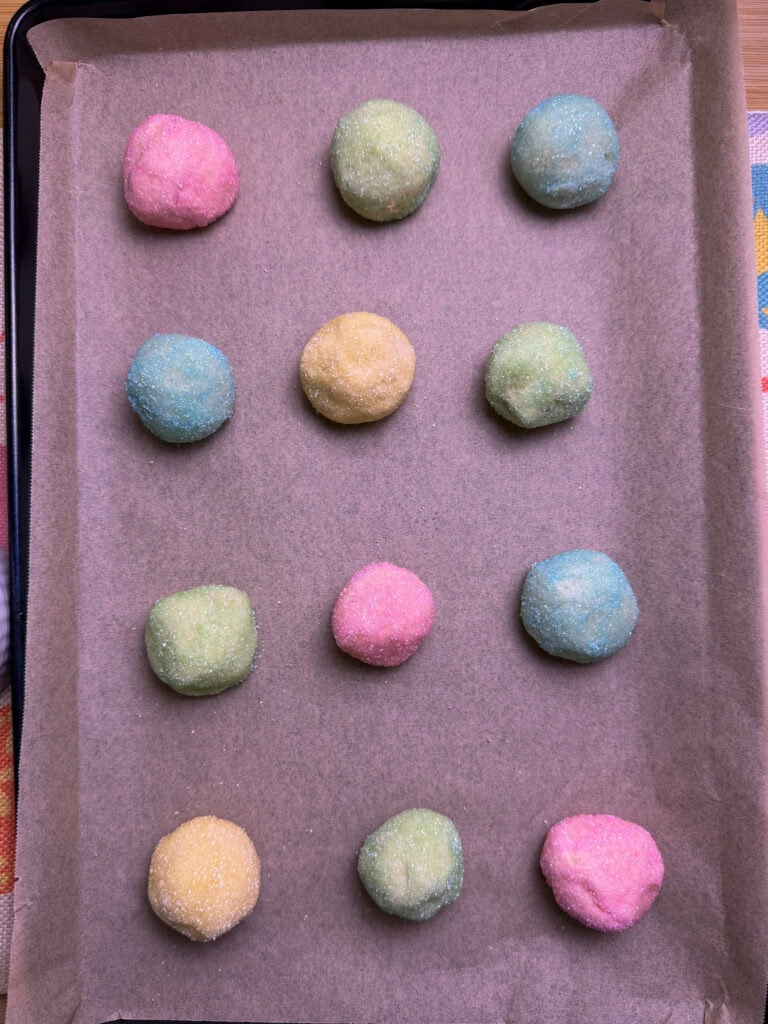 A baking tray lined with parchment paper holds twelve dough balls in pastel colors—pink, yellow, blue, and green—arranged neatly in a grid pattern, ready to be baked for an Overnight Hashbrown Breakfast Casserole treat.