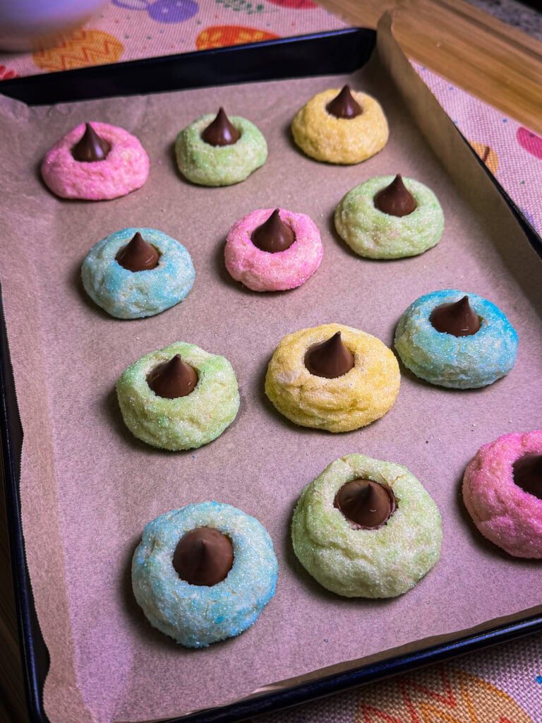 A baking tray lined with parchment paper holds colorful sugar cookies, each topped with a chocolate kiss—perfect alongside an Overnight Hashbrown Breakfast Casserole for a cheerful brunch spread. The cookies are pastel pink, green, yellow, and blue.