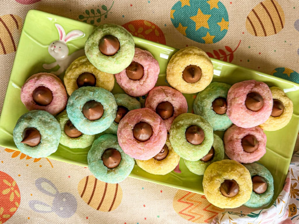 A rectangular plate with pastel-colored cookies topped with chocolate kisses, set on a tablecloth decorated with Easter eggs and bunny patterns. A bunny figurine decorates the plate’s edge—perfect next to your Overnight Hashbrown Breakfast Casserole.